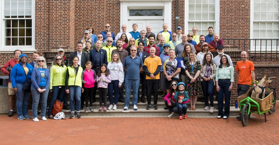The whole group of adults and children standing on the steps of one of the U of D buildings, and a dog sits in a parked cargo bike